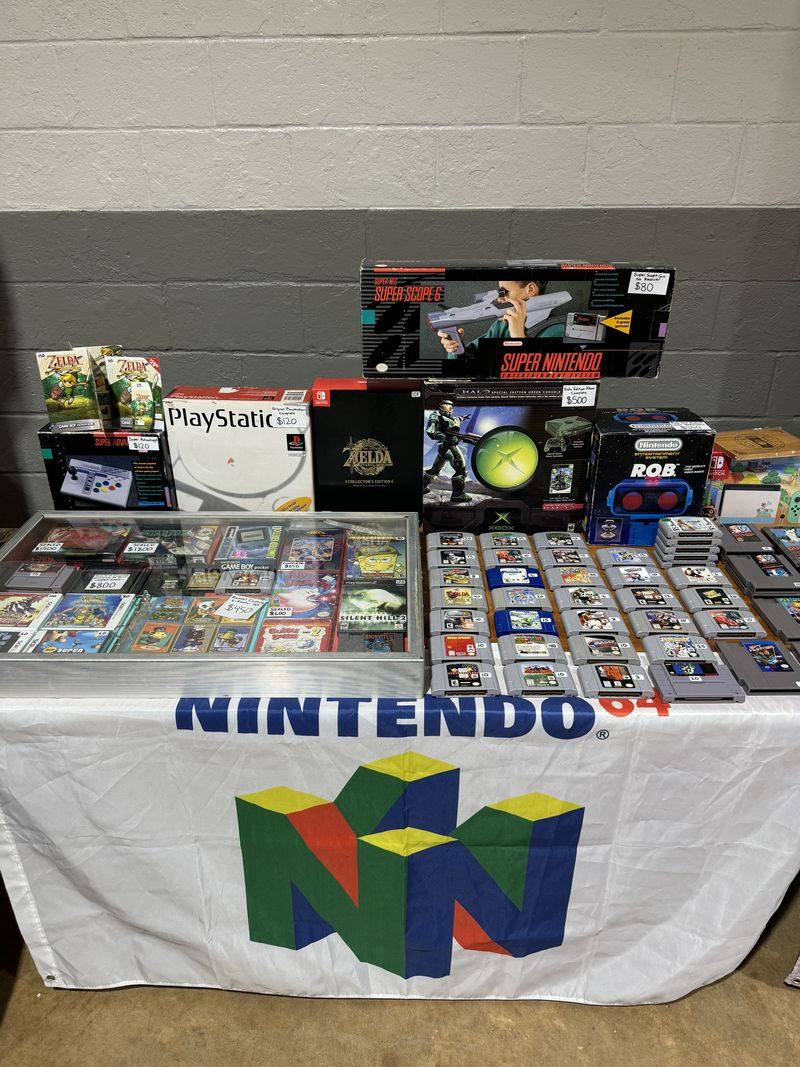 A retro gaming dealer's table with boxed Super Nintendo, PlayStation, and Xbox consoles above rows of Nintendo 64 cartridges