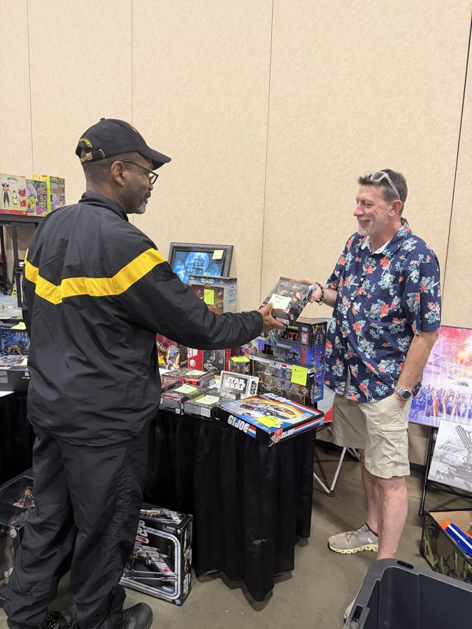 A smiling vendor handing a boxed collectible to a customer over a table of G.I. Joe and Star Wars toys