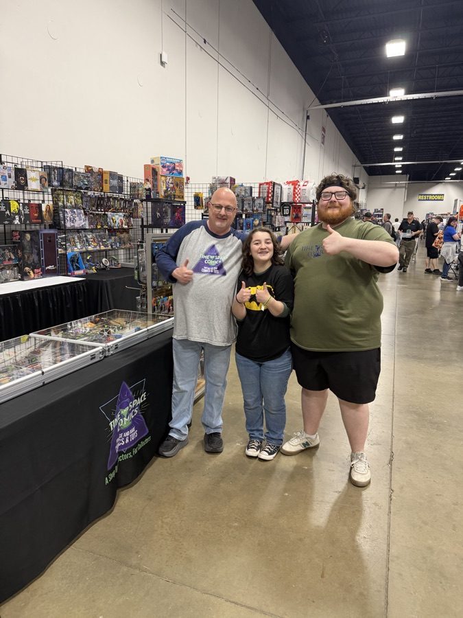 A comic shop crew giving thumbs-up in front of vintage comics and action figures